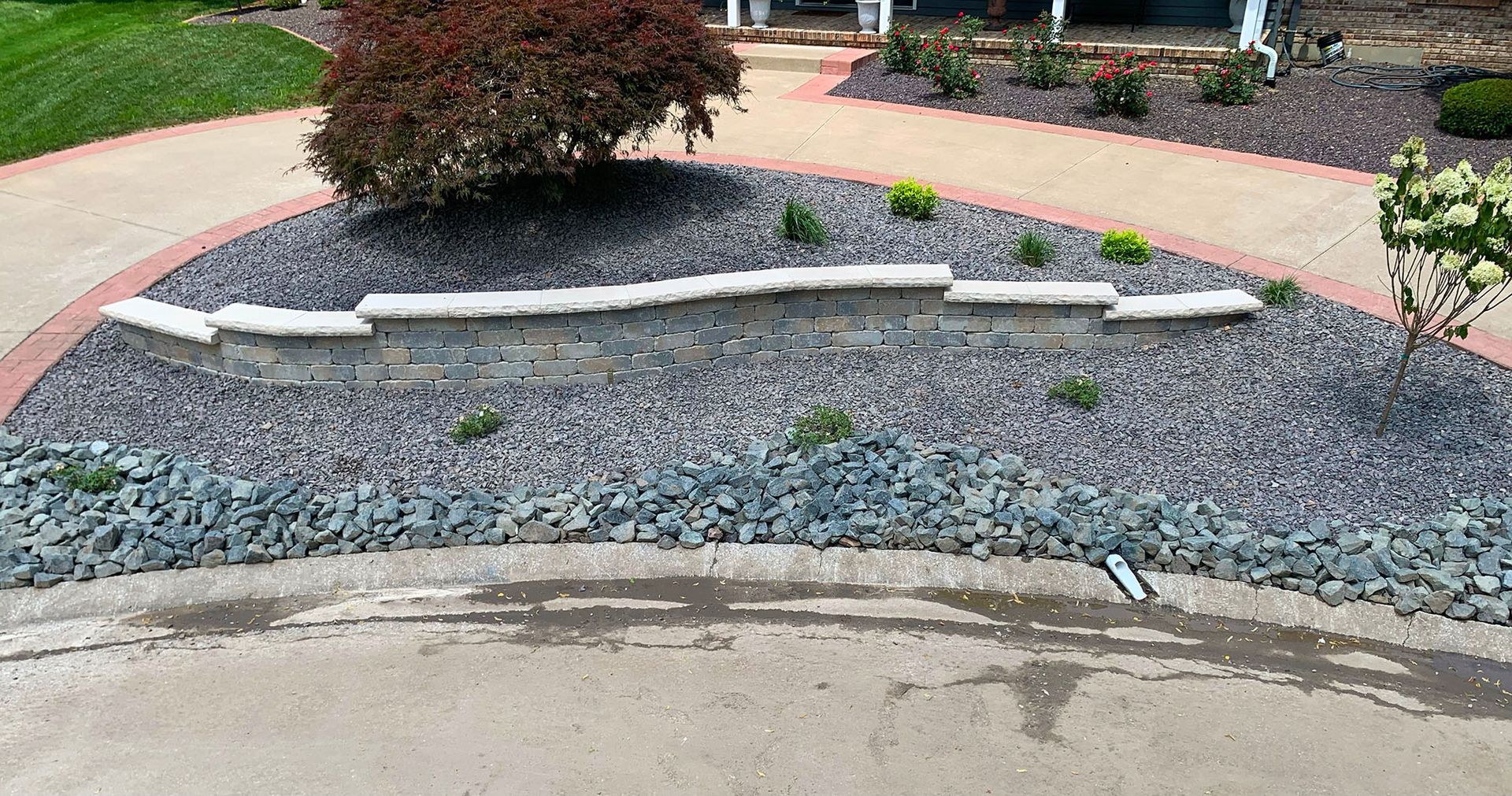 A curved stone wall sits in a landscaped garden bed with grey gravel and shrubs, bordered by a concrete walkway.
