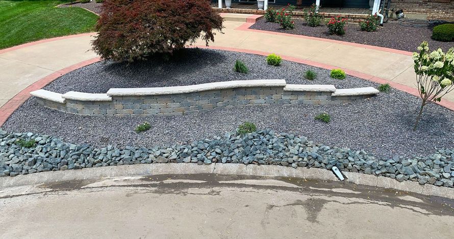 A curved stone wall sits in a landscaped garden bed with grey gravel and shrubs, bordered by a concrete walkway.