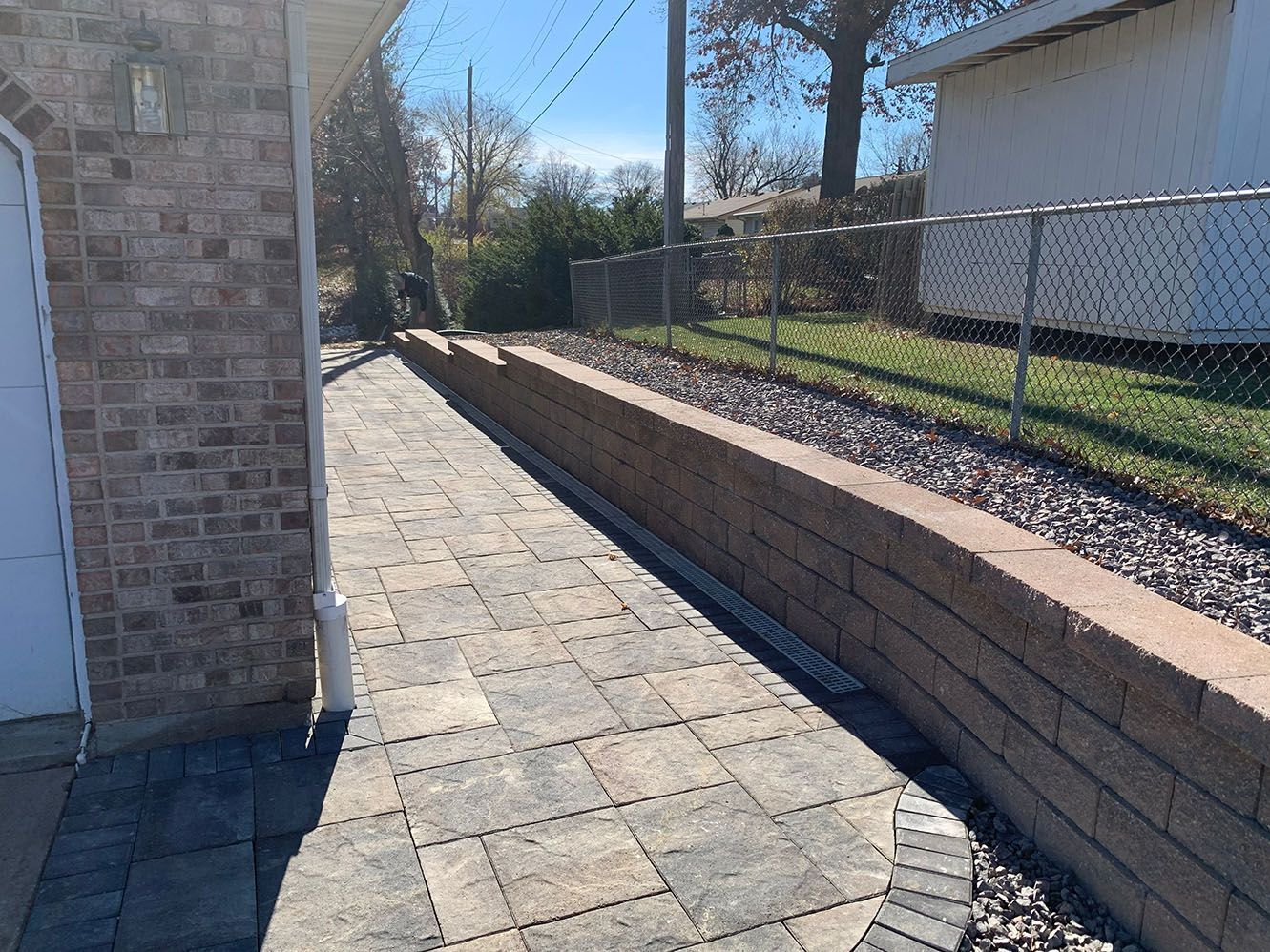 A stone patio walkway runs alongside a terraced retaining wall made of stacked blocks, bordering a gravel bed and fence.