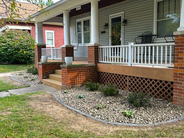 A gray house with a covered front porch, brick columns, white railings, and a gravel garden bed in the front yard.