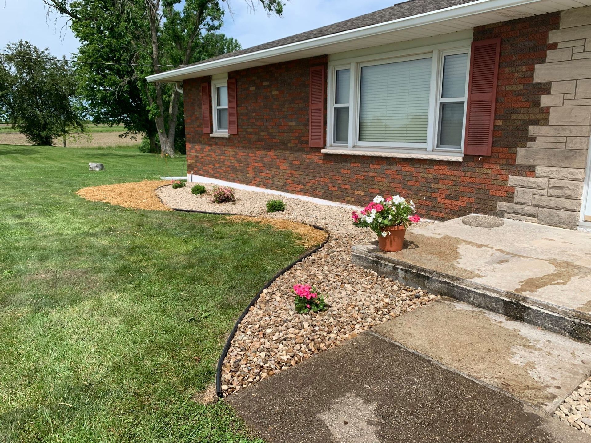 A landscaped flower bed with brown mulch and light-colored gravel edges a brick house next to a green lawn.