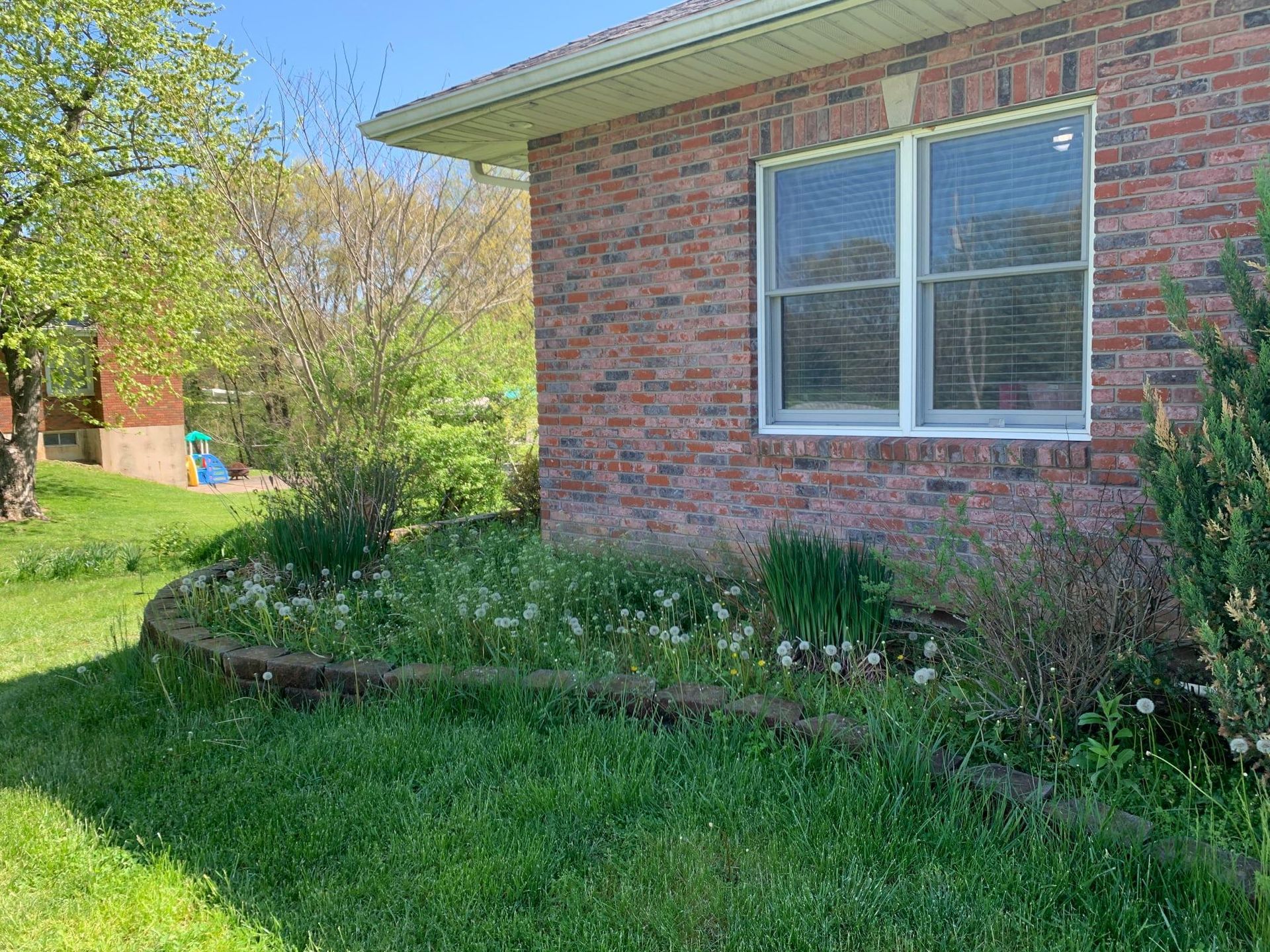 A brick house corner with a curved stone garden bed featuring green plants and white flowers on a sunny day.
