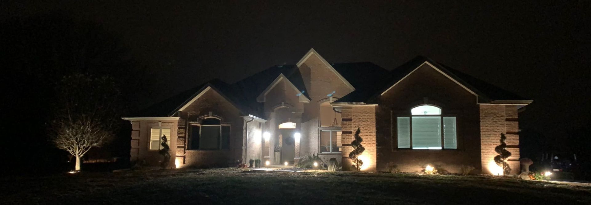 A brick house at night illuminated by exterior landscaping lights, with a dark sky in the background.