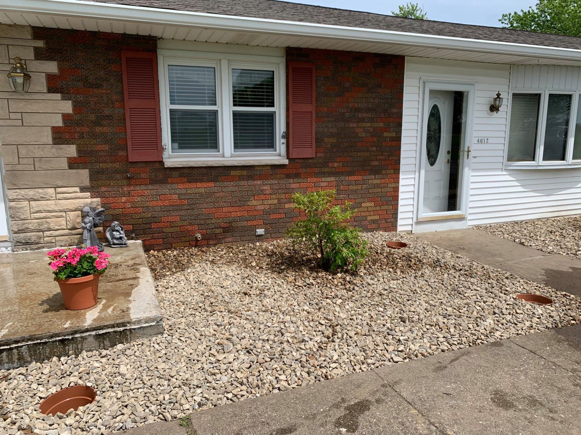 A house exterior with brick and white siding, a front door, two windows with red shutters, and a gravel front yard.
