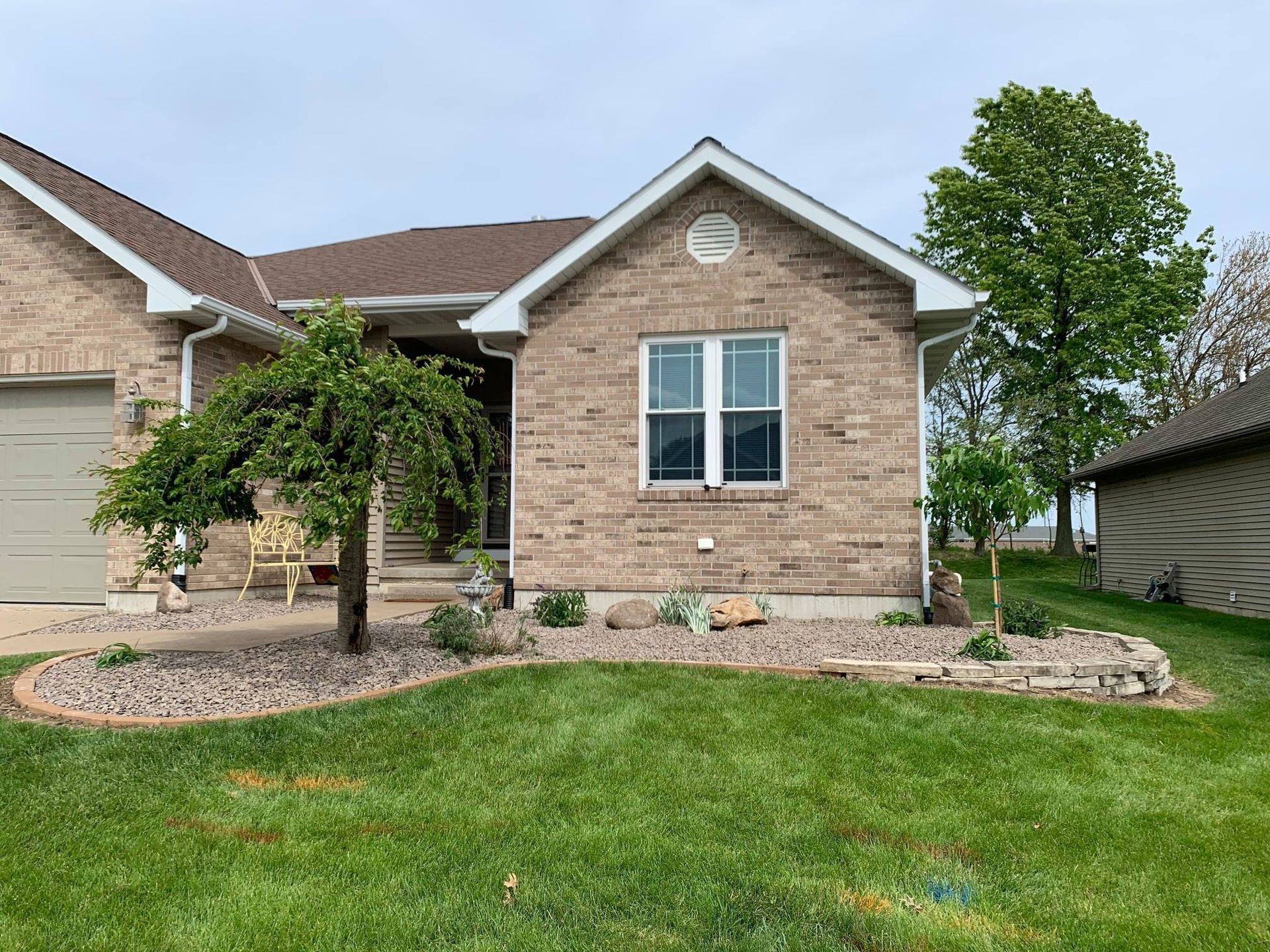 Tan brick house with a front lawn, a decorative stone-bordered flower bed, and a small weeping tree.