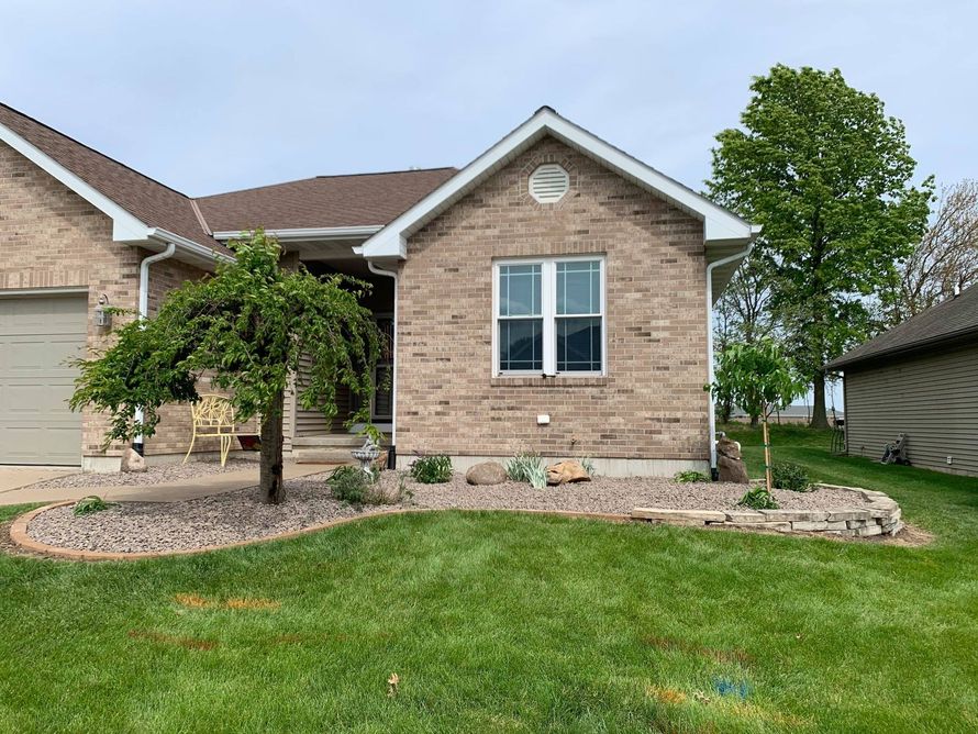 A tan brick house with a front landscaping bed featuring a small weeping tree, river rock, and a stone border.