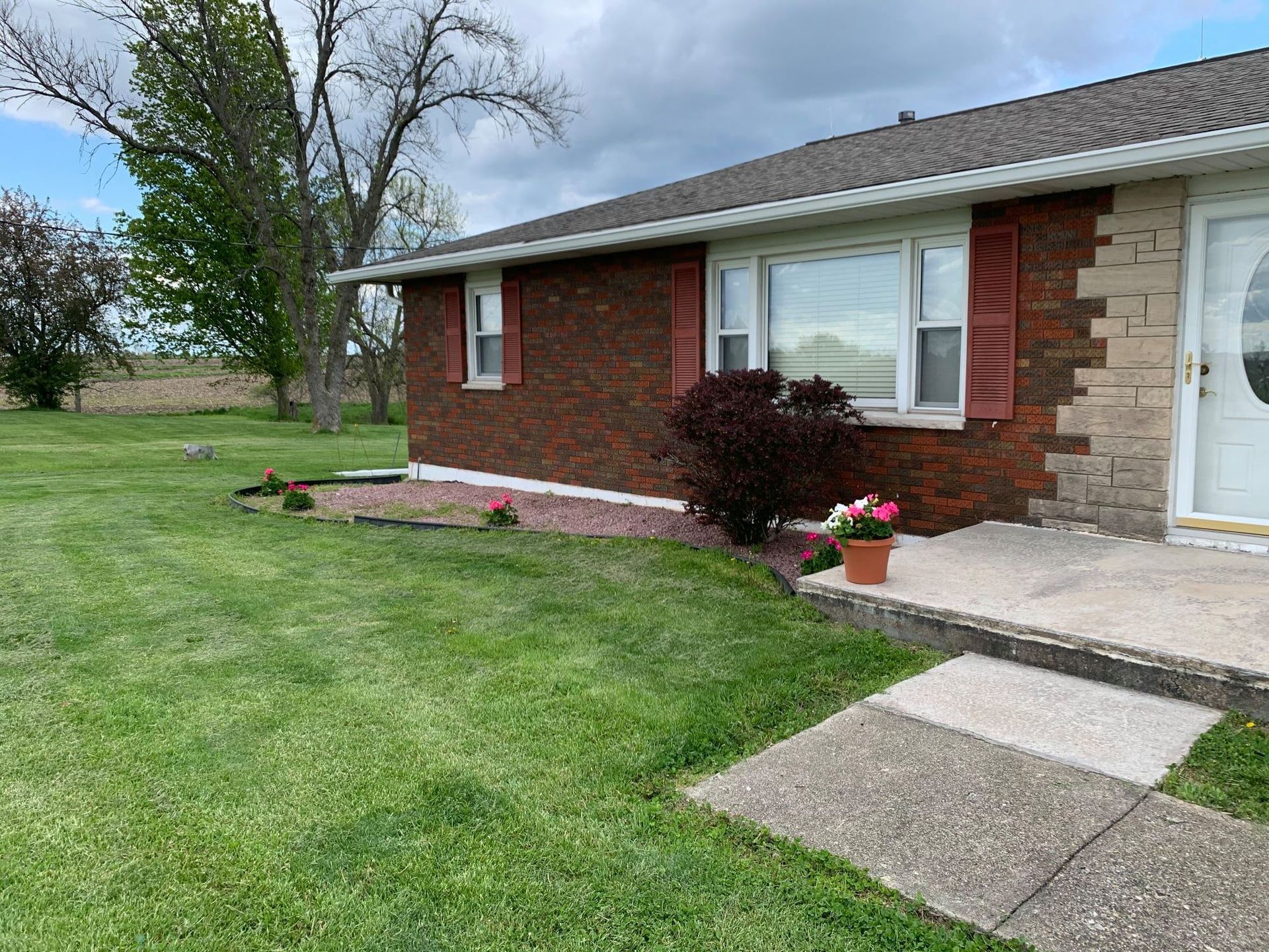 A red brick house with a white door and shutters, featuring a garden bed with a potted flower on a concrete porch.