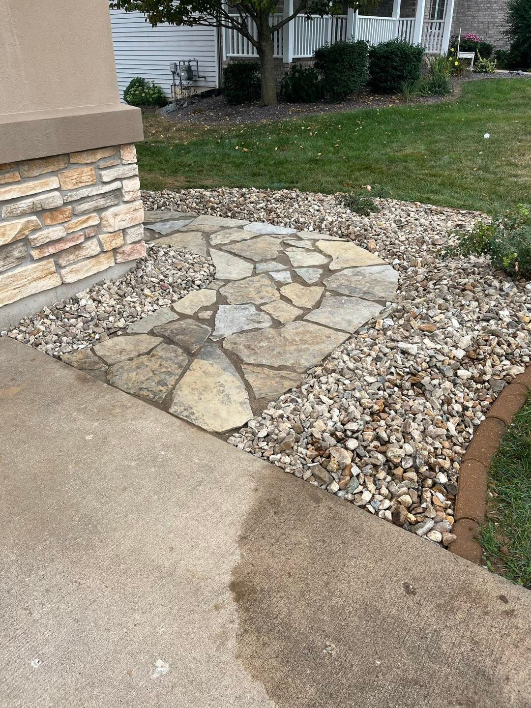 A flagstone walkway segment embedded in gravel next to a concrete sidewalk and a stone-faced house wall.