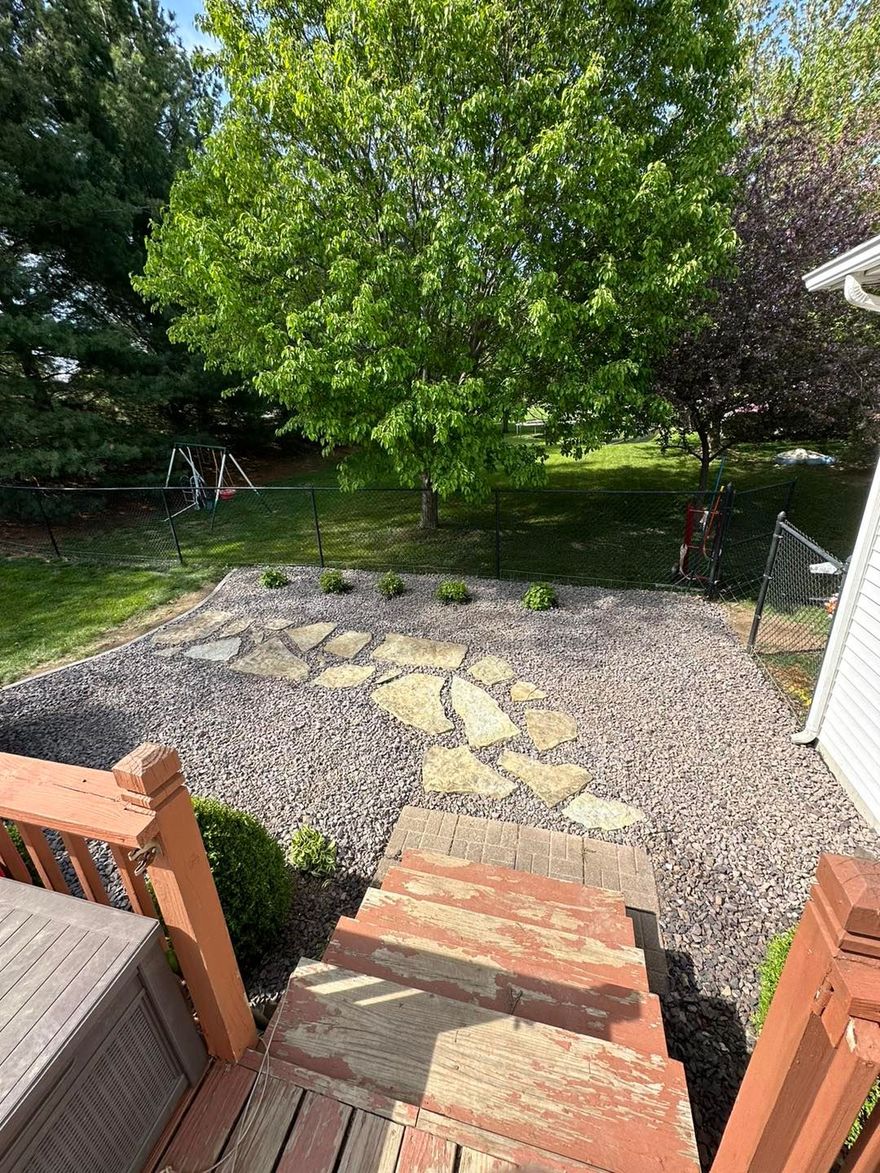 A stone path set in grey gravel leads from a wooden deck into a green backyard with a large tree.