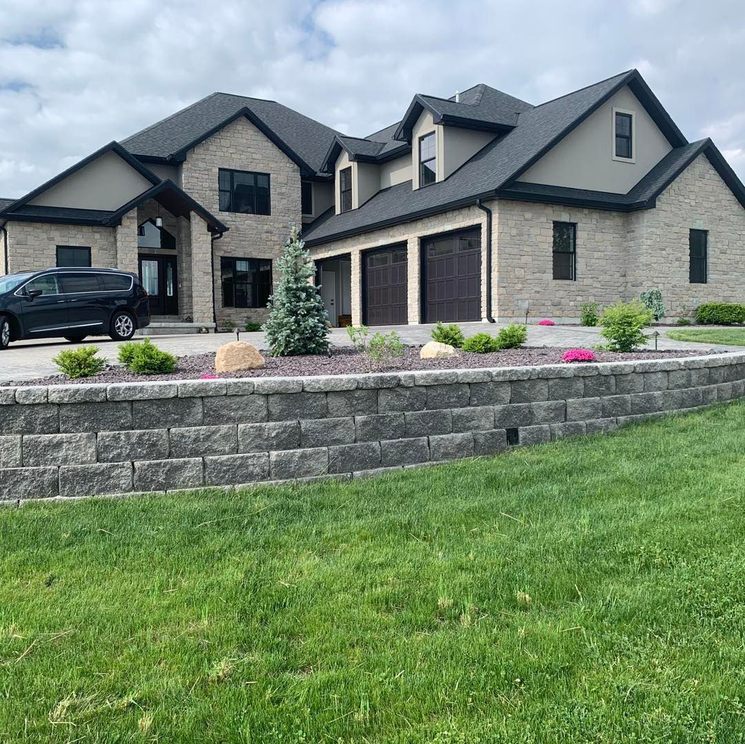 A tan brick multi-story suburban house with a dark roof and three-car garage, fronted by a stone retaining wall and lawn.