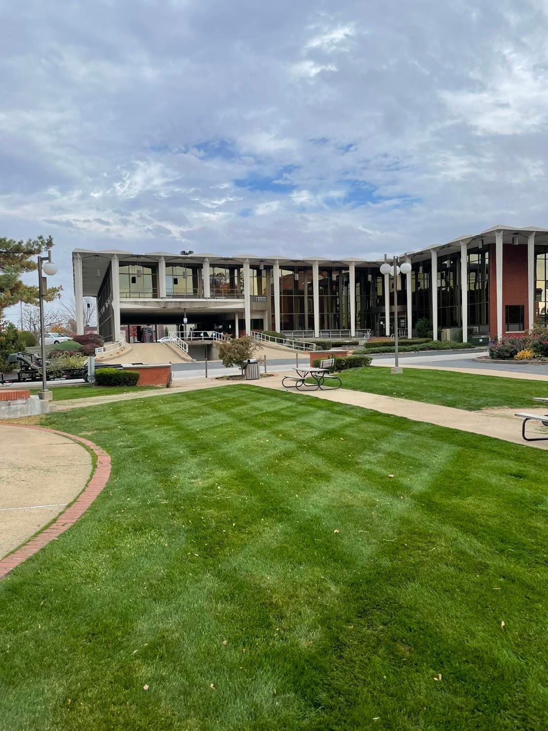 A two-story building with glass walls and pillars stands behind a large, mowed lawn under a cloudy sky.