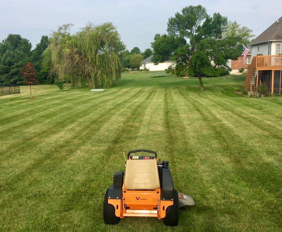 A bright orange stand-on zero-turn mower parked on a freshly mown suburban lawn with distinct stripes.