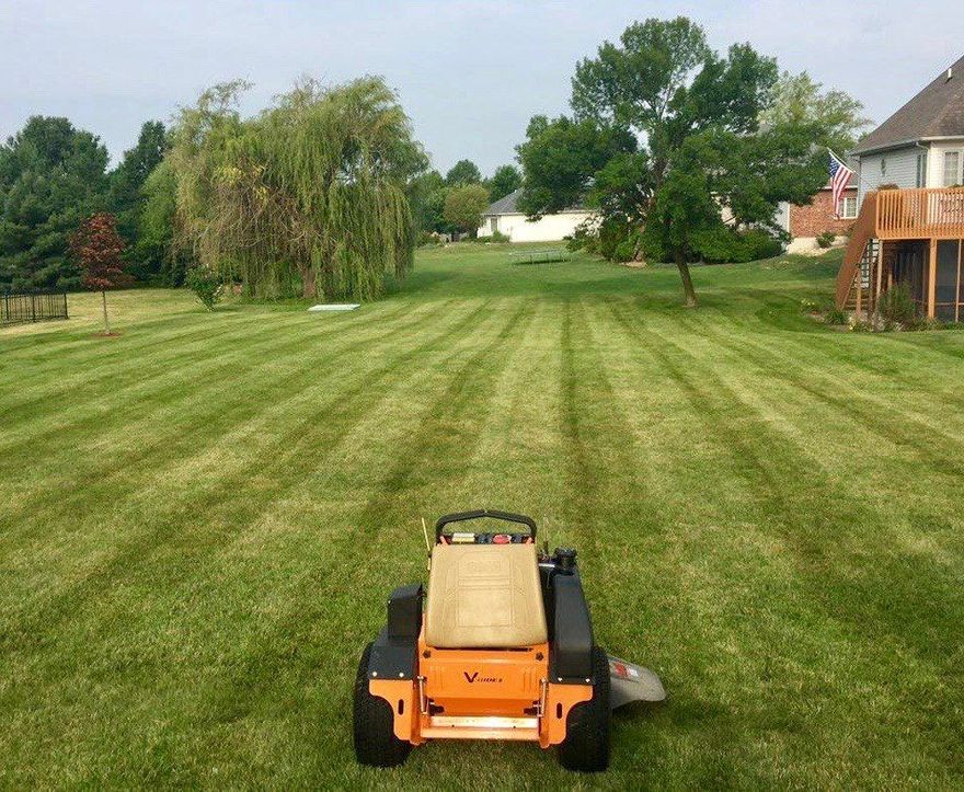 An orange zero-turn mower sits on a neatly striped residential lawn with trees and a house in the background.