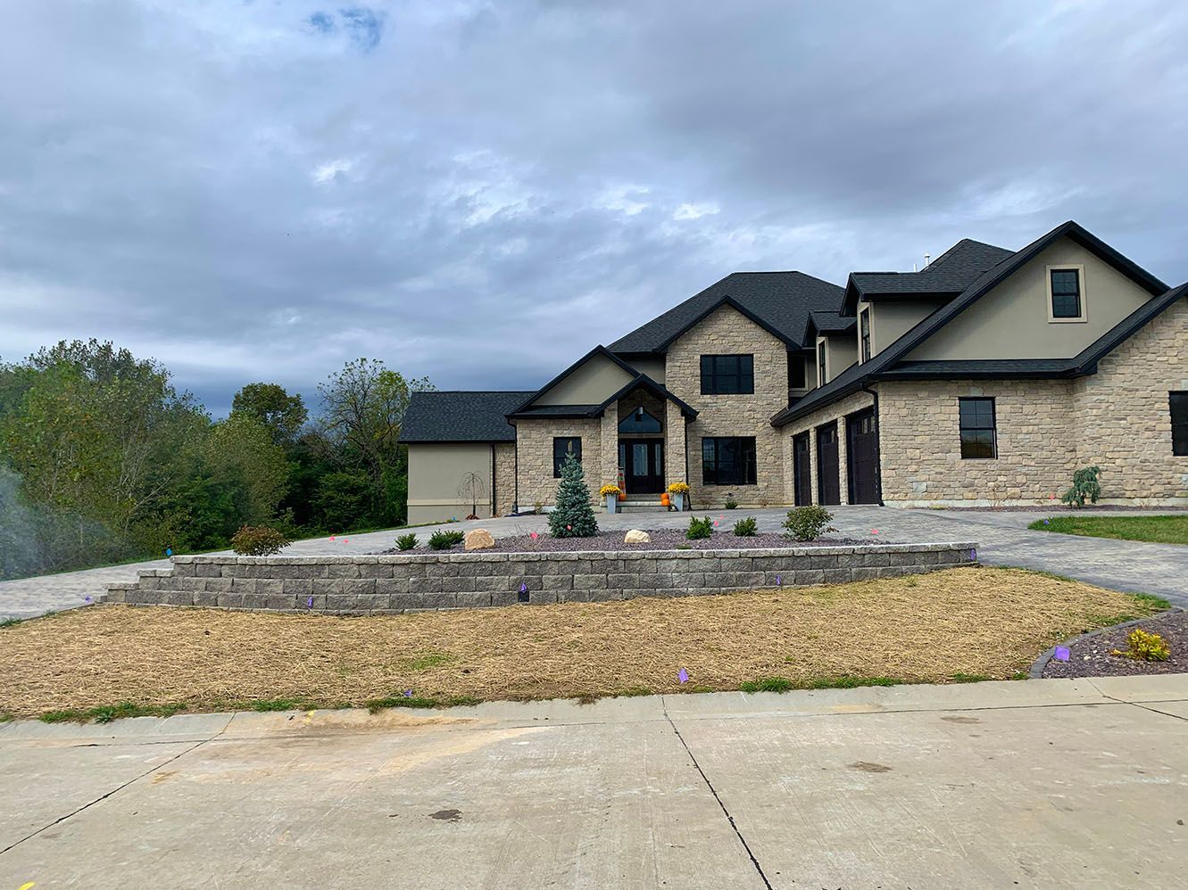 A large two-story stone house with a dark roof, a concrete driveway, and a landscaped front yard under a cloudy sky.