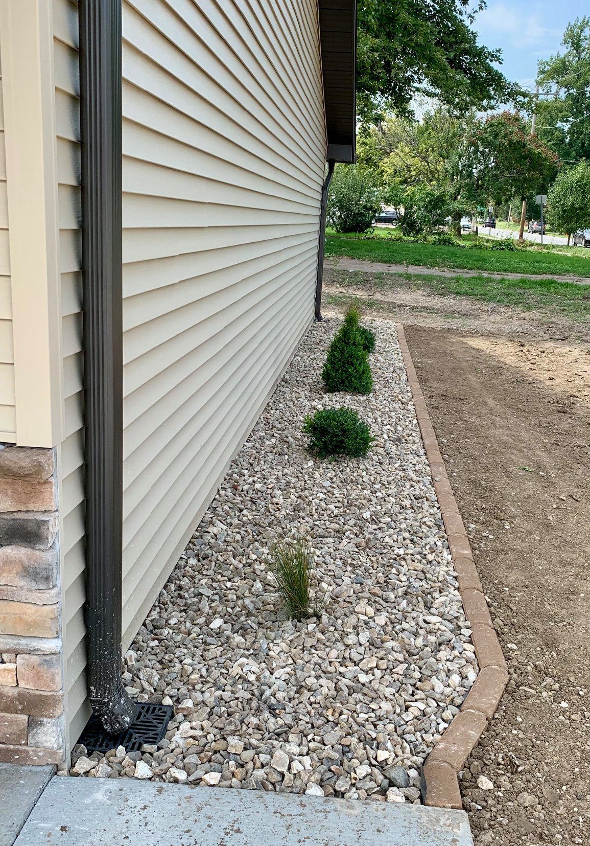 A side view of a house exterior with light siding, a downspout, a gravel flower bed, and small evergreen bushes.