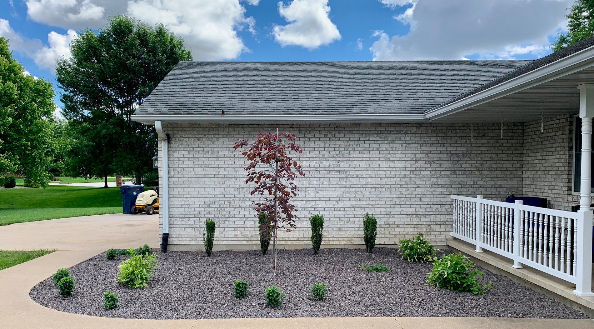 A white brick house exterior with a small front garden featuring dark gravel, a young red tree, and small shrubs.