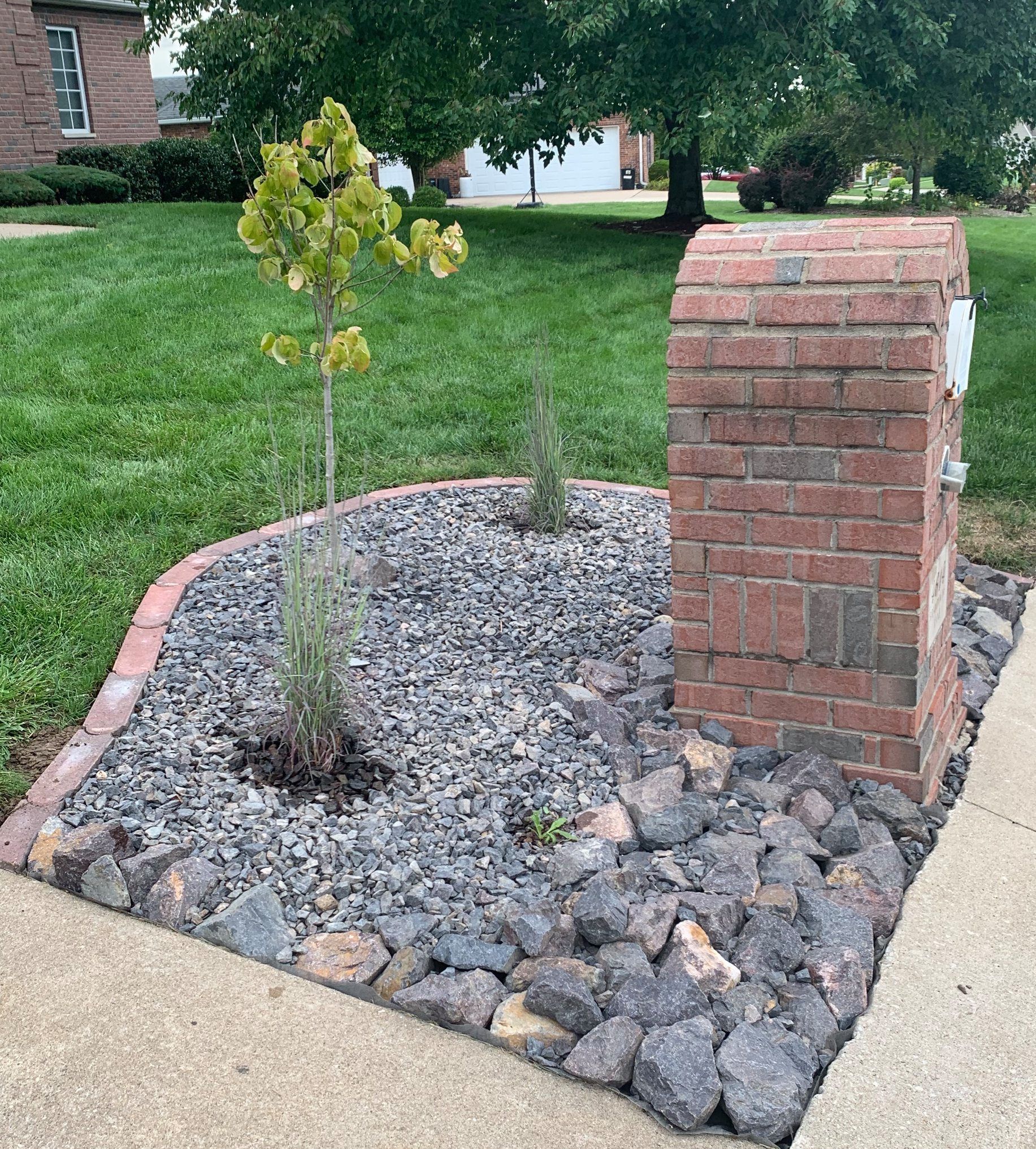 A brick mailbox stands in a landscaped bed of gray gravel and large stones next to a sidewalk and green lawn.