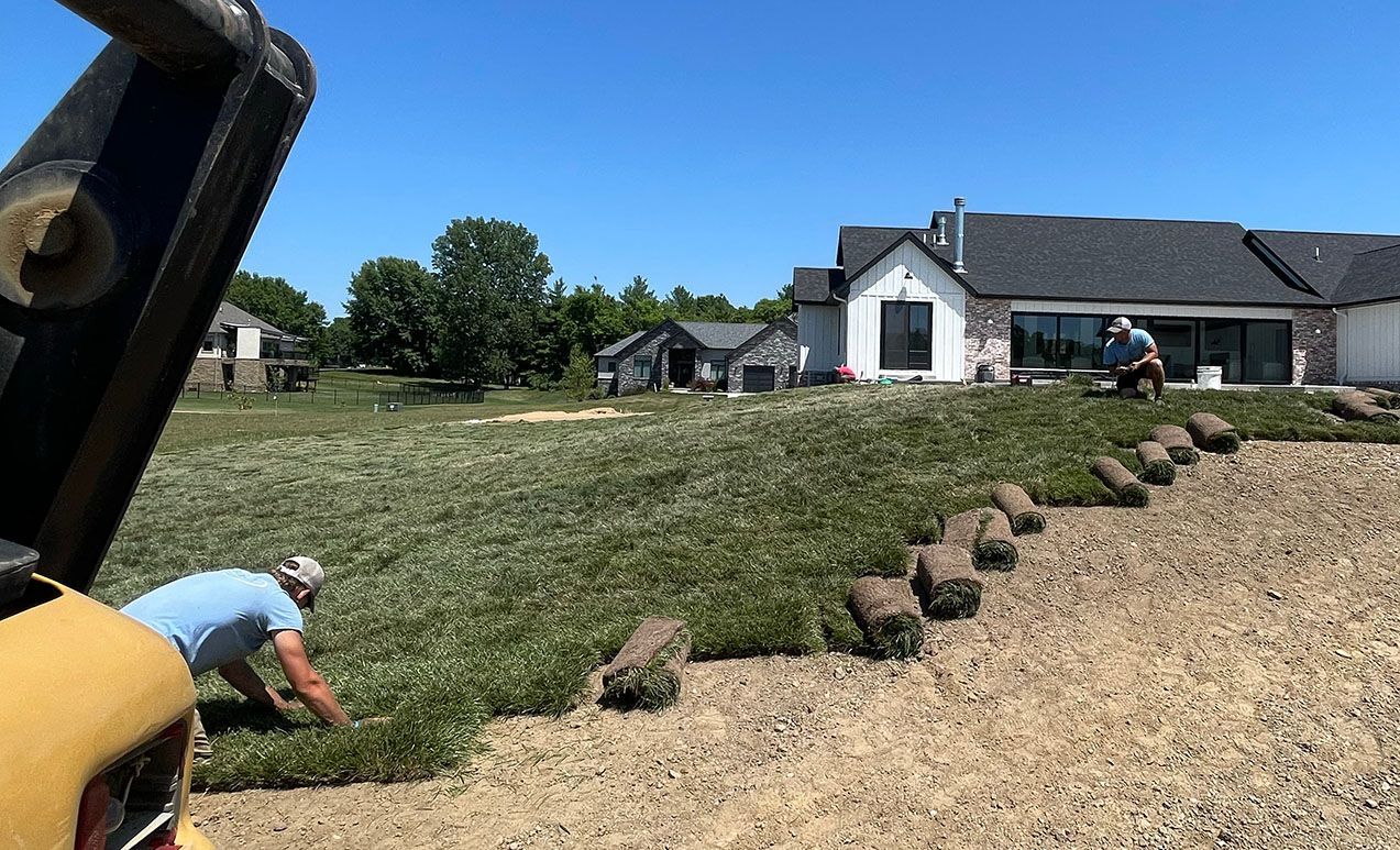 Two people lay rolls of green sod on a grassy residential lawn near a new house on a sunny day.