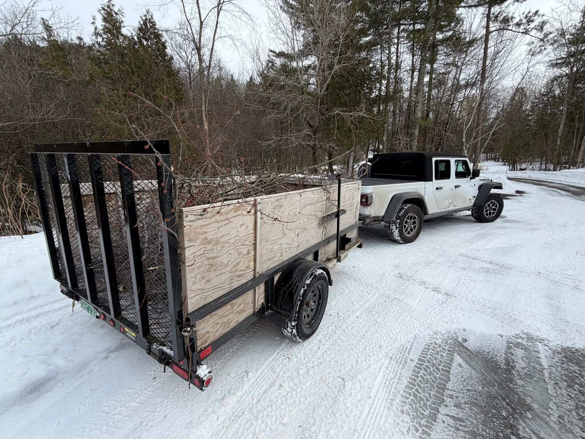 White Jeep towing a small black trailer on a snowy road beside winter trees