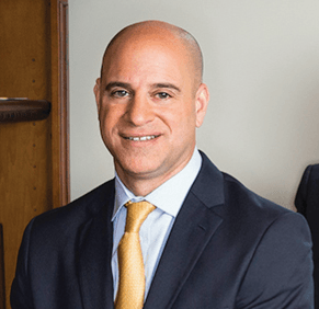 Man in dark suit, light blue shirt, and gold tie smiles at the camera.