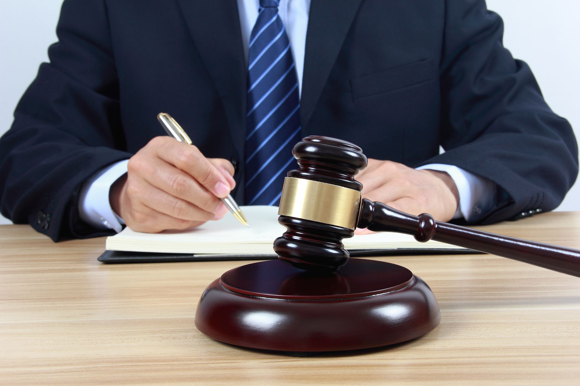 Judge’s gavel placed on a wooden desk with a person writing in a notebook in the background. Judge’s gavel placed on a wooden desk with a person writing in a notebook in the background.