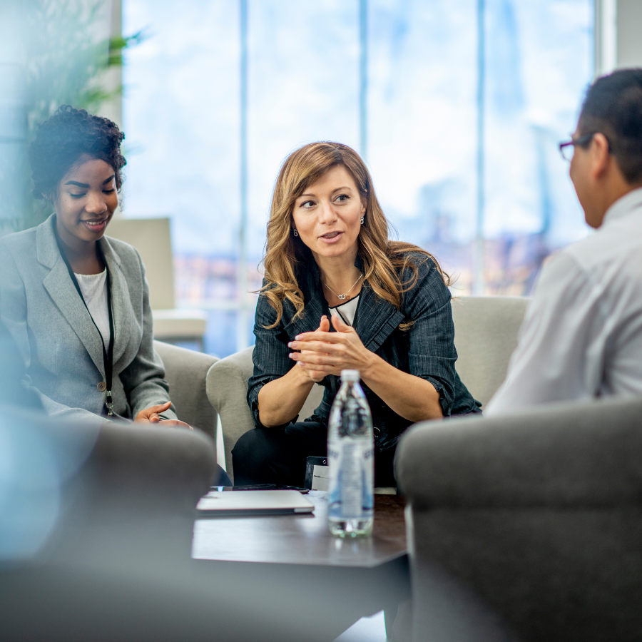 Three coworkers in a meeting, one speaking, seated around a table in a bright office