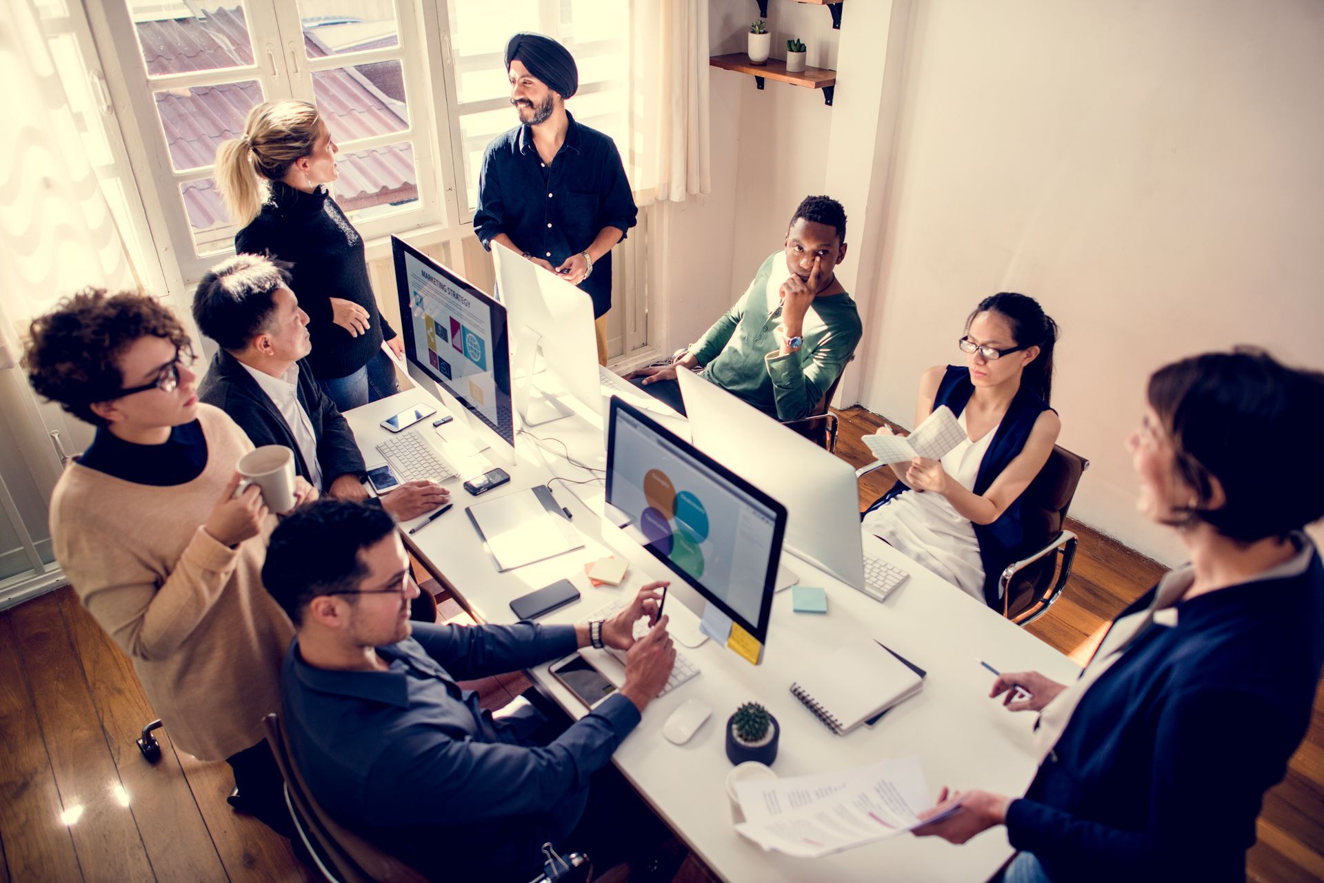 Team meeting around a long white conference table with desktop computers in a bright office.