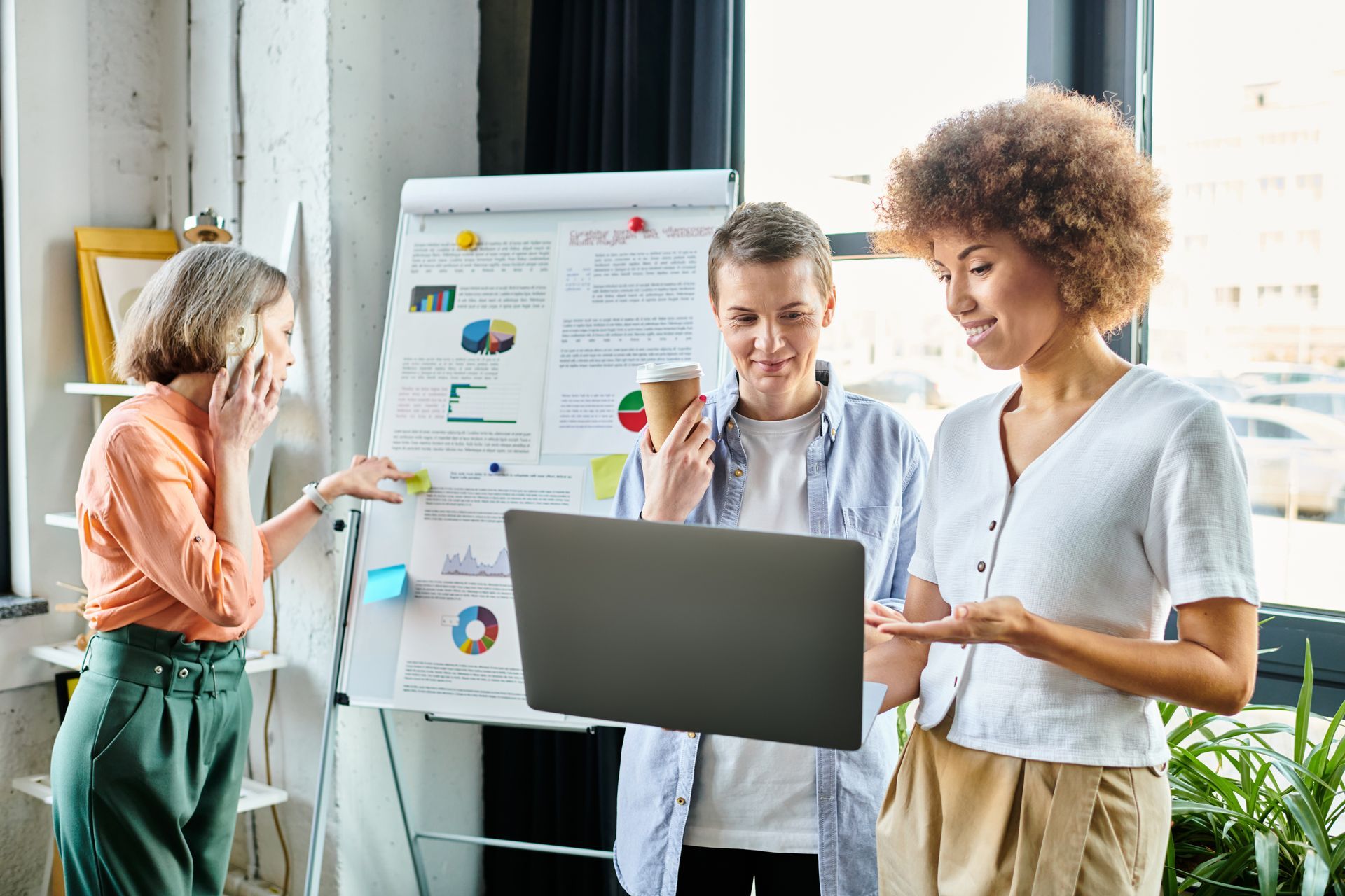 Three coworkers discussing a presentation beside a laptop and chart board in a bright office