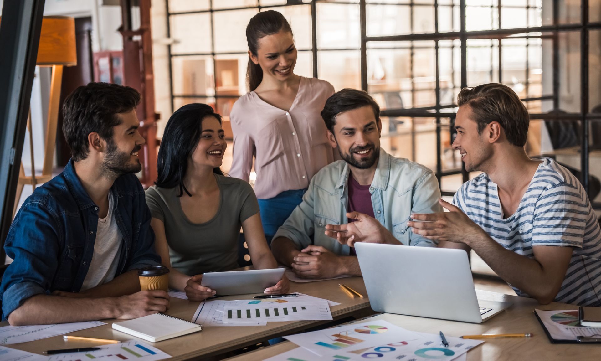 Five coworkers meeting around a table with a laptop and documents in a bright office