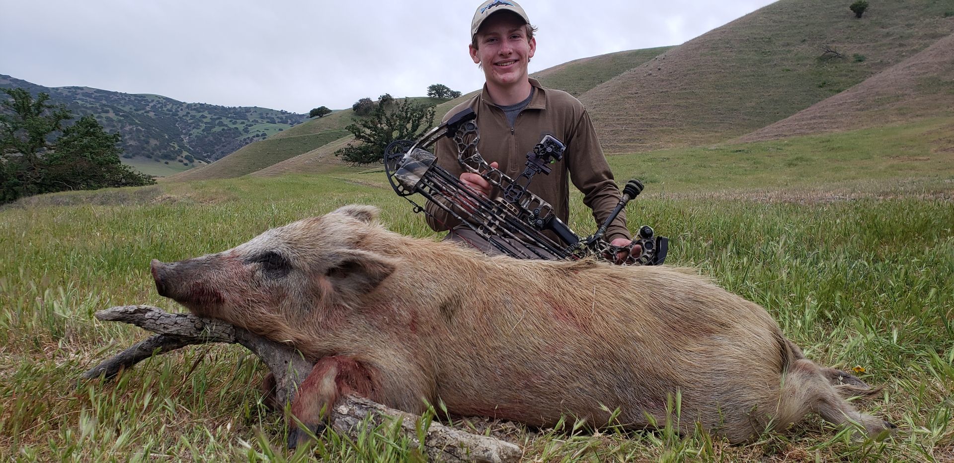 Hunter standing behind a large wild boar in a grassy field with hills in the background