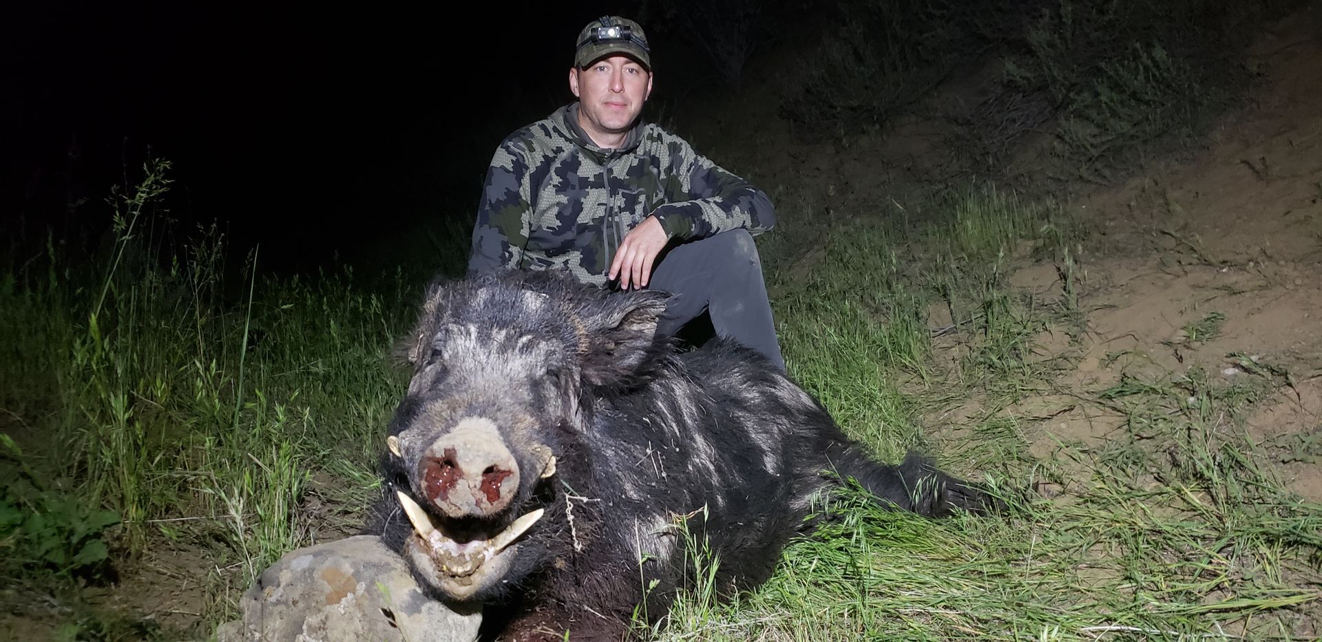 Person crouches beside a dead wild boar at night, illuminated by a bright flash in a grassy area.