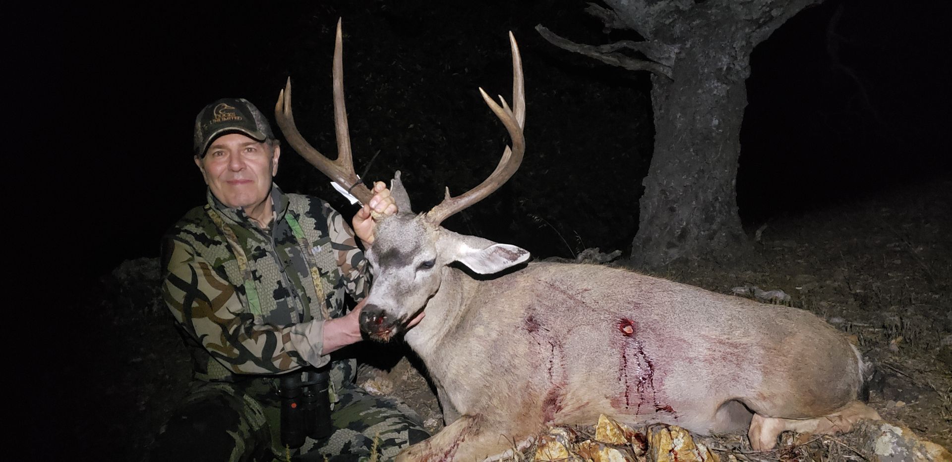 Hunter posing with a large deer at night by a tree