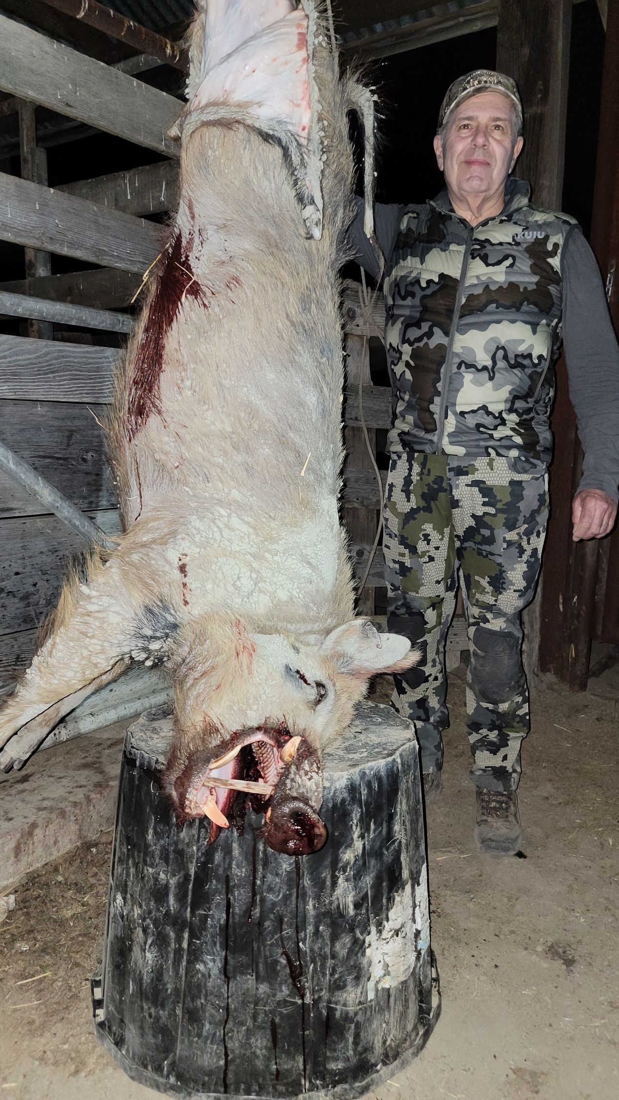 Person in camouflage stands beside a hanging deer carcass in a dim shed