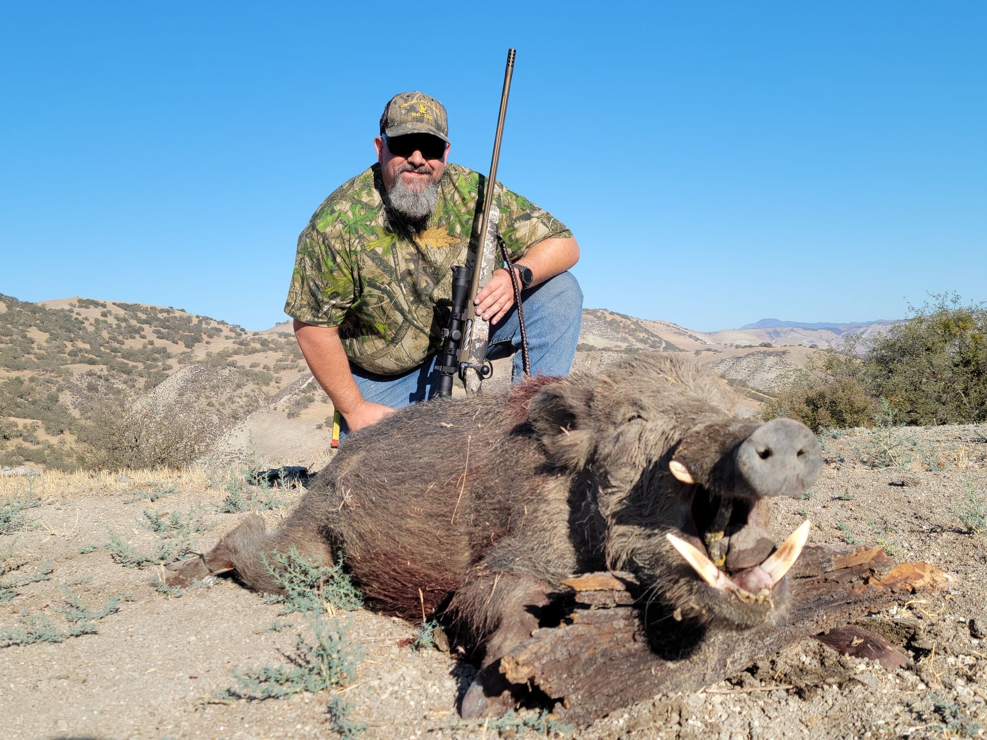 Hunter kneeling beside a dead wild boar in a dry desert landscape under a clear blue sky