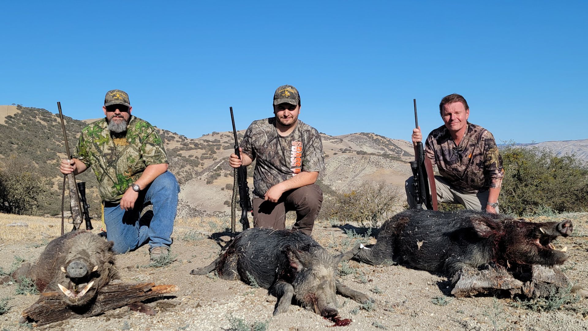 Three hunters kneel with rifles beside two wild boars in a dry, rocky landscape.