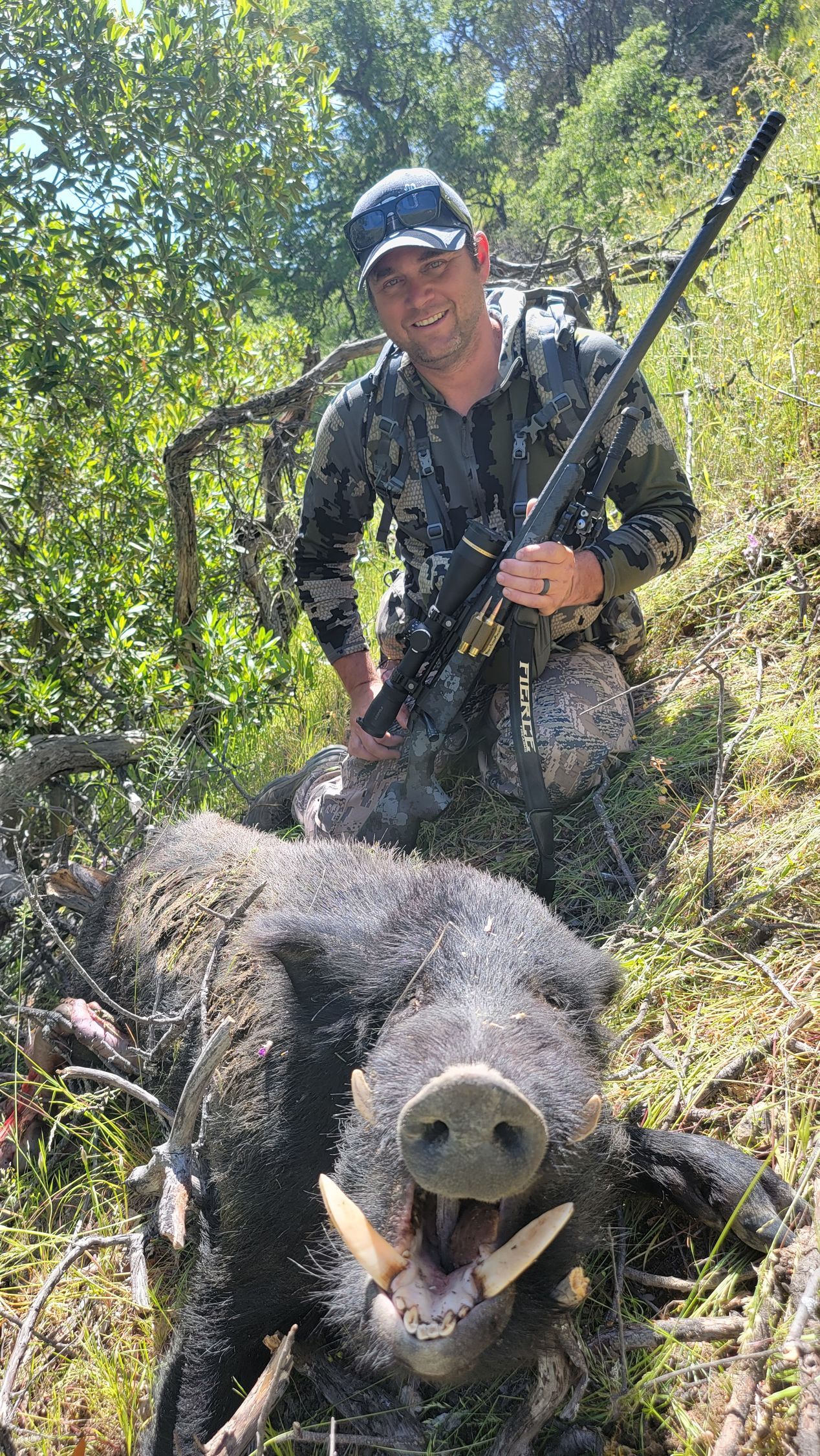 Person in camouflage kneels behind a wild boar in a wooded area, holding a rifle.