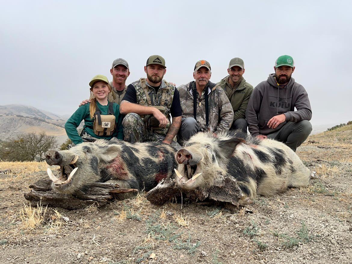 Five hunters kneel behind two wild boars in a dry field under an overcast sky.