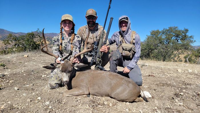 Three hunters kneel behind a large wild boar in a dry field, holding rifles and smiling.