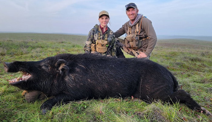 Two hunters kneel behind a large black bear in a grassy field under a cloudy sky