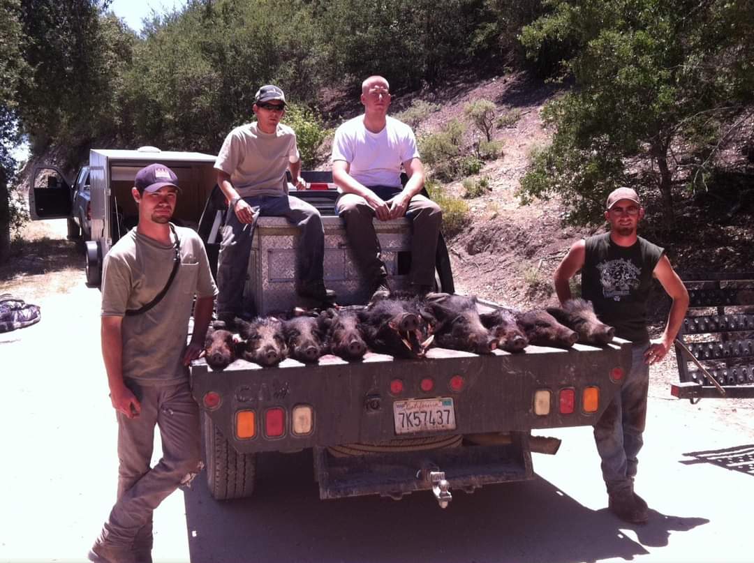 Four people posing on the back of a truck loaded with wild boar, outdoors on a dirt road