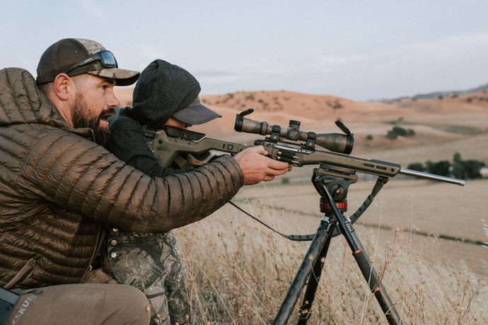 Two people aiming a scoped rifle from a tripod in a dry field at dusk