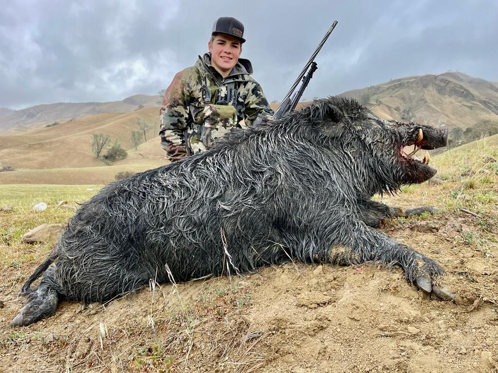 Hunter kneels beside a large wild boar on a grassy hillside with mountains in the background