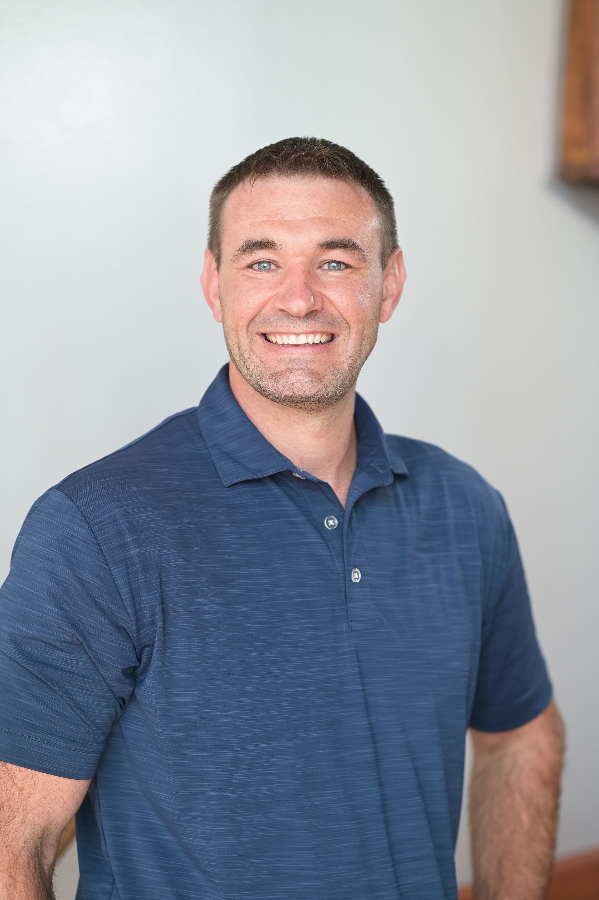 Man with short brown hair and a blue polo shirt smiling, standing in front of a light gray wall.
