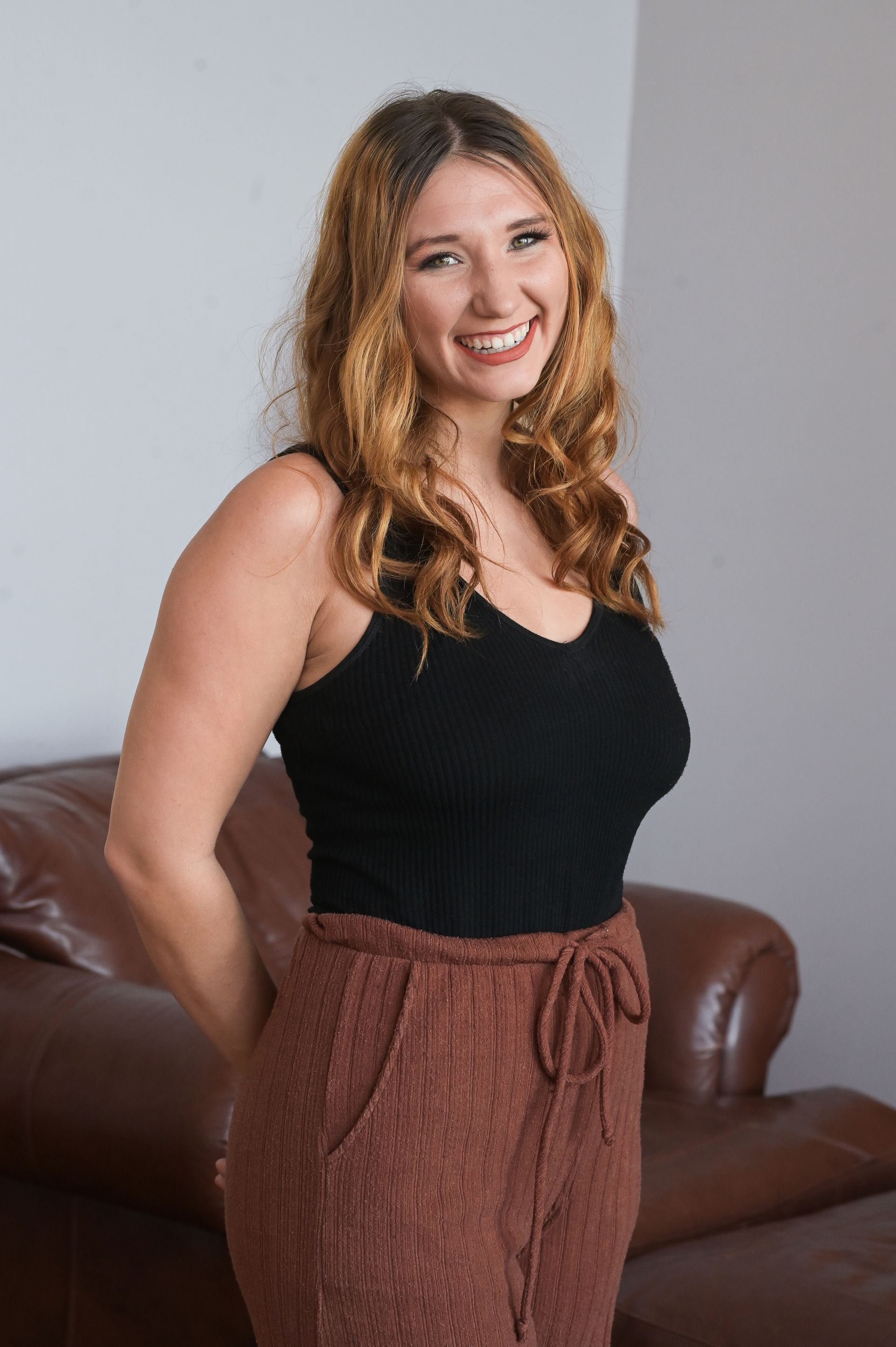 Woman with long wavy auburn hair smiles, wearing a black tank top and brown drawstring pants, posing in front of a brown leather sofa.
