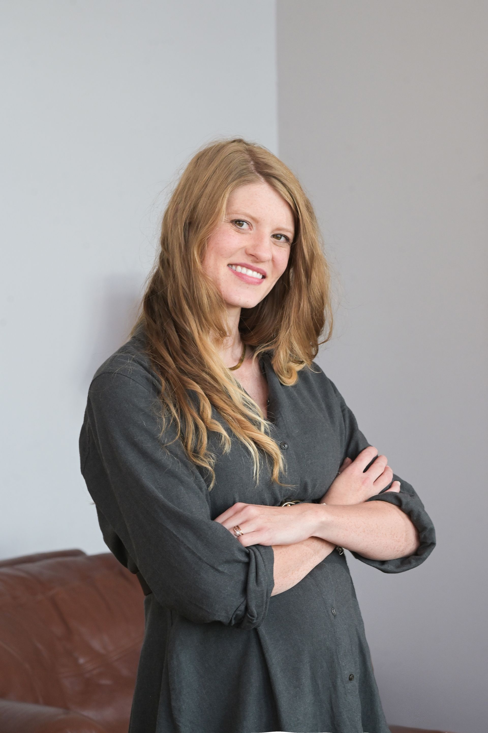 A woman in a grey dress is standing with her arms crossed and smiling.