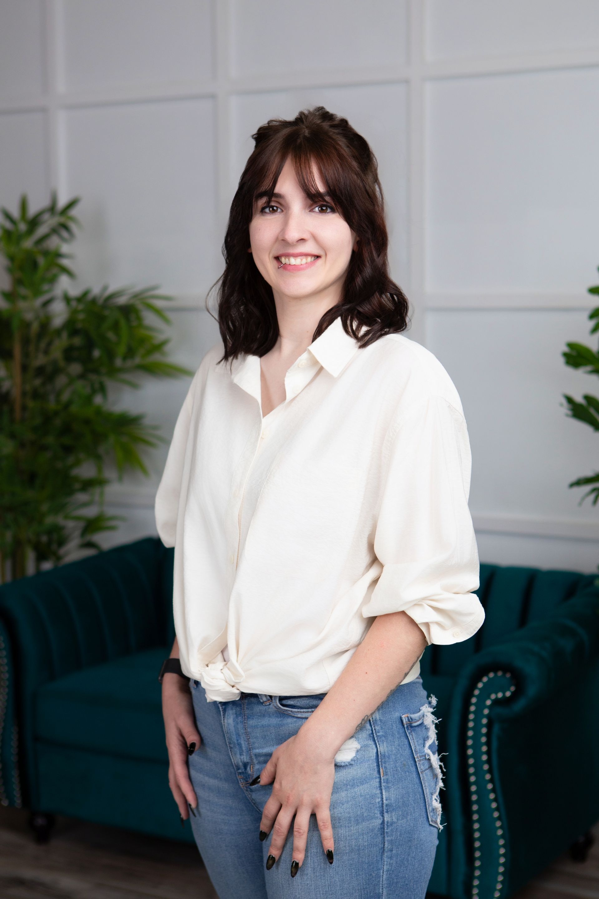 Woman with dark hair and bangs smiles in front of a teal sofa, wearing a white shirt and jeans.