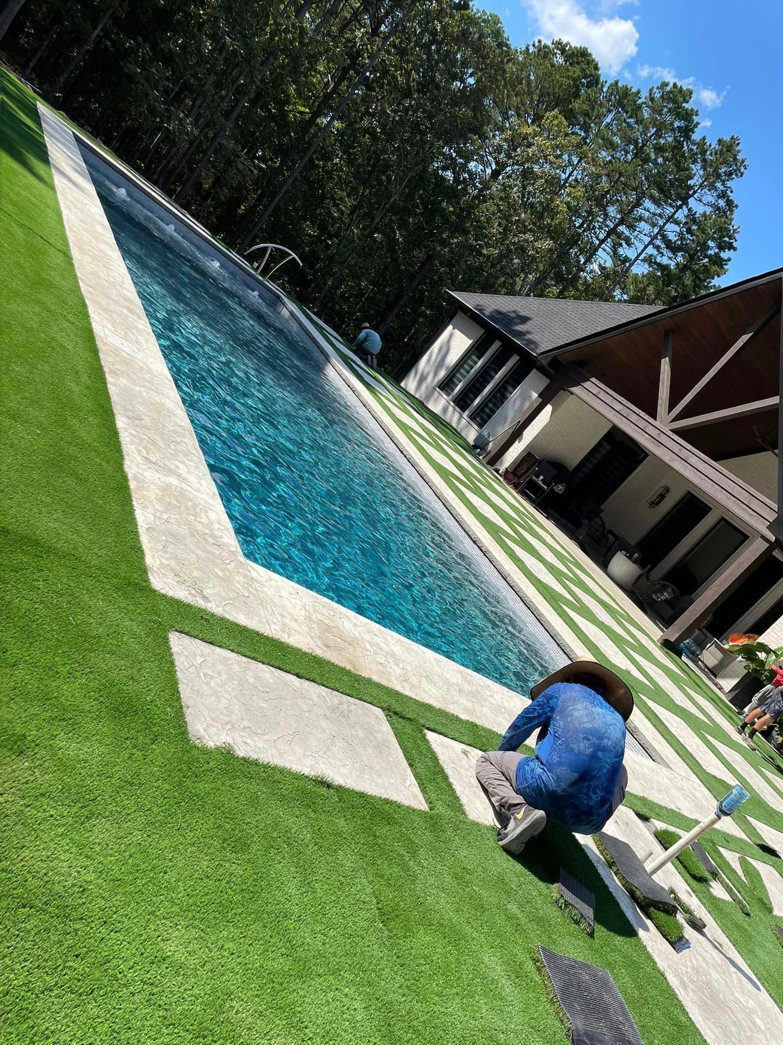 A man is kneeling on the grass next to a swimming pool.