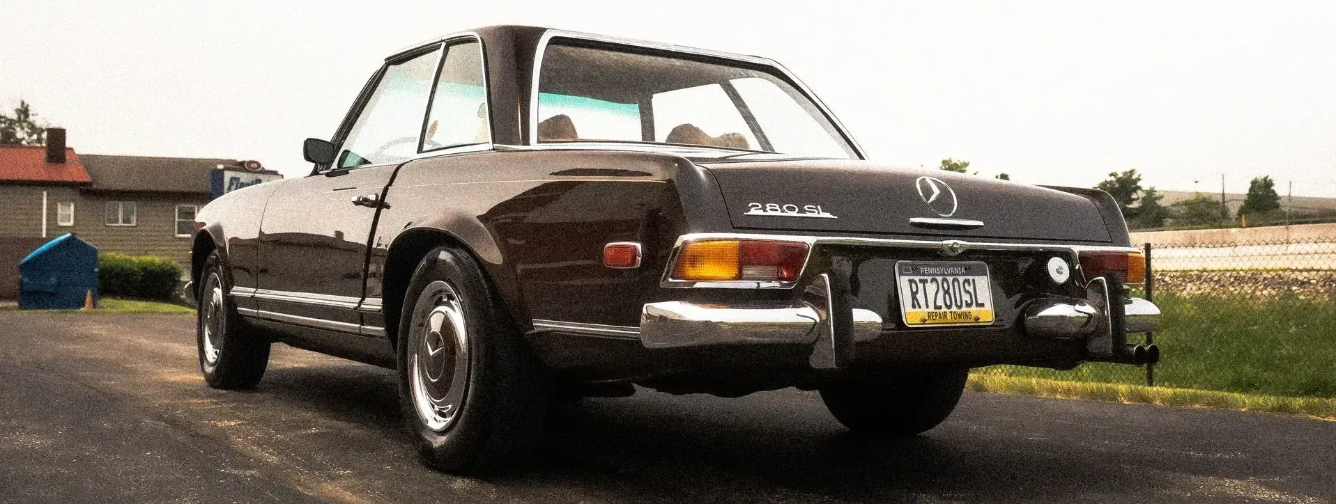 Dark brown Mercedes-Benz SL, viewed from behind, parked on a paved surface. A rectangular hardtop is in place.