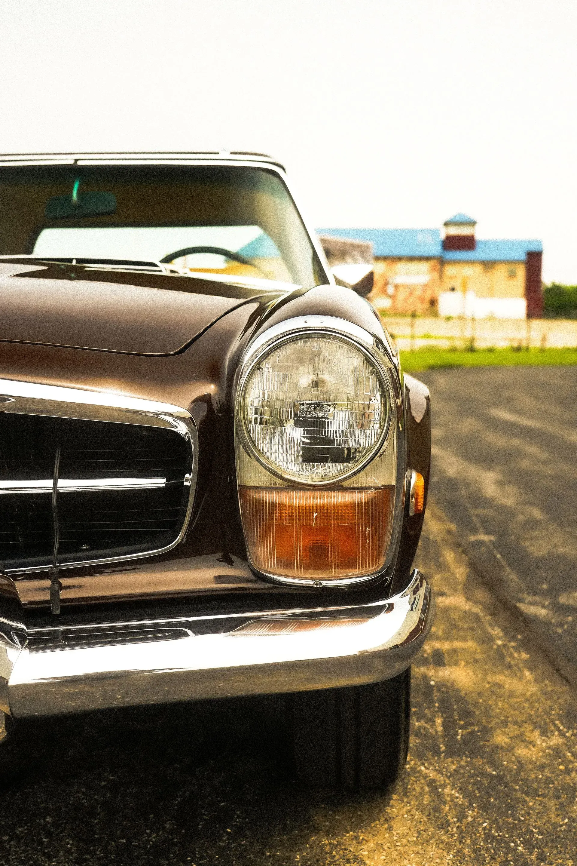 Brown classic car, close-up of headlight and grill. Chrome bumper. Building in the background.