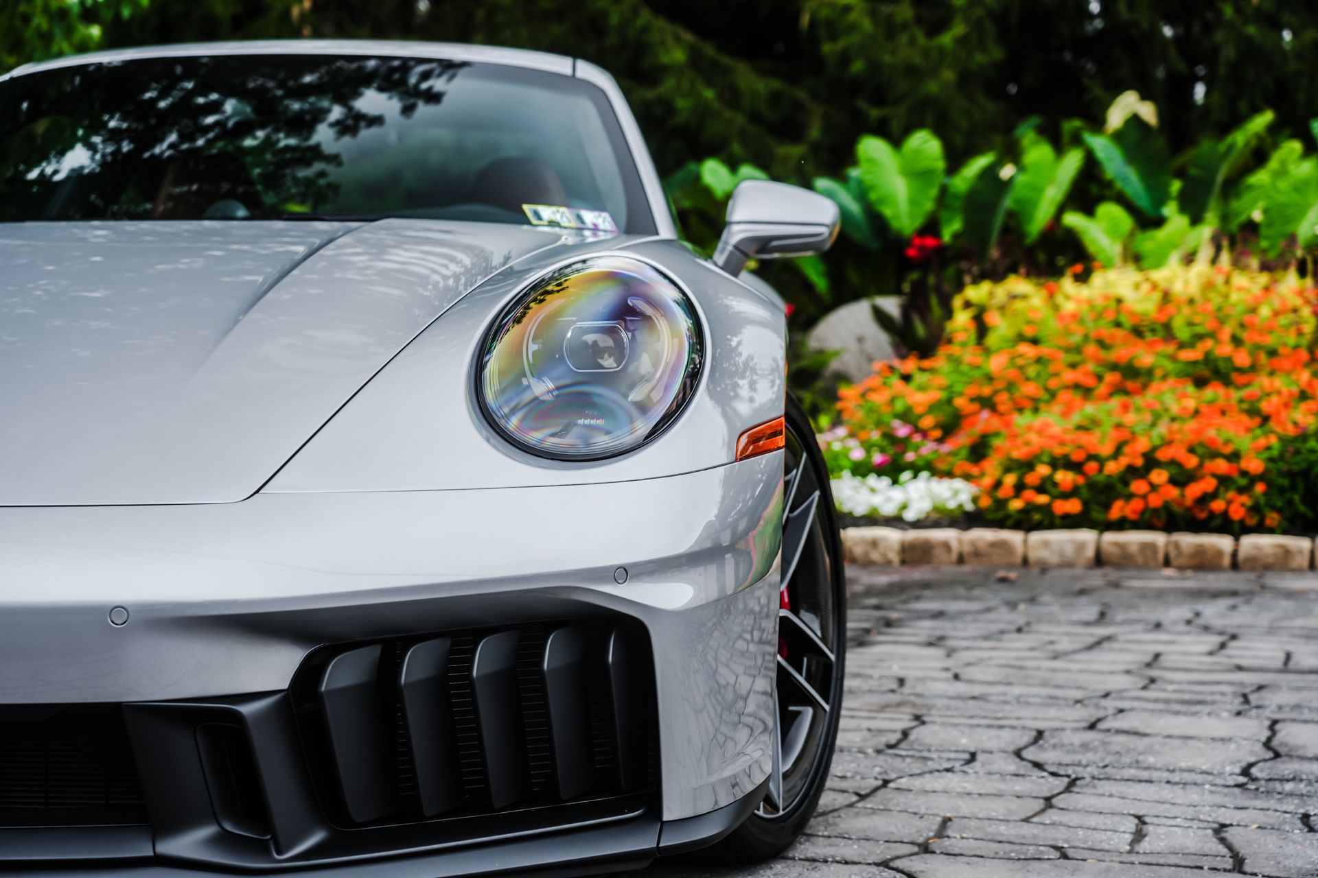 Silver Porsche sports car parked on a stone driveway, with a colorful flower garden in the background.